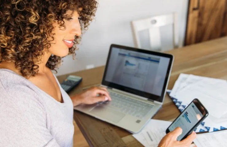 Woman managing finances at home using smartphone and laptop, with bills on the table.