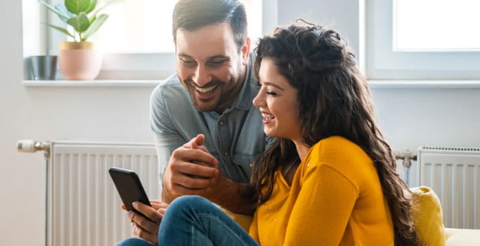 Happy couple sitting together and looking at a smartphone using digital banking in a cozy living room.