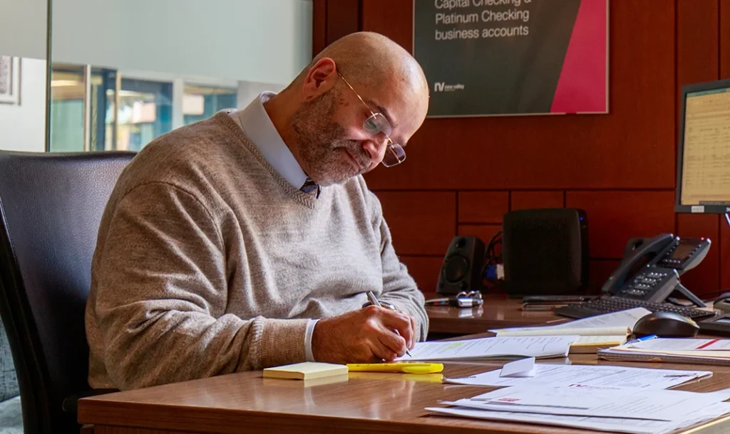 Bank employee writing notes at a desk with paperwork and a phone nearby, inside a New Valley Bank & Trust branch.
